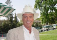 Older light skinned man with white hair smiles wearing a white wide brimmed hat, white button down and checkered blazer