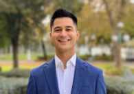 A man in a blue blazer and white dress shirt stands smiling at the camera