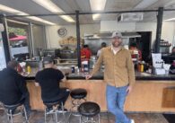 A man stands inside a diner, leaning on the counter next to customers