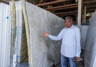 An older man in a button down shirt and jeans stands gesturing to slabs of granite next to him