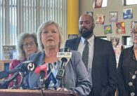 A woman speaks into microphones at a podium, with two women and a man standing behind her