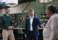 Three man standing together, with the man in the center pointing toward the gut of the man standing to the right.