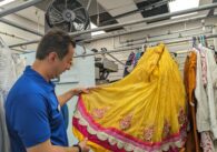 A middle-aged man in a blue work shirt holds up a yellow and pink sequinned Indian sari.