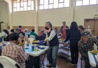 An older man passes out plates of food to older adults seated at a table.