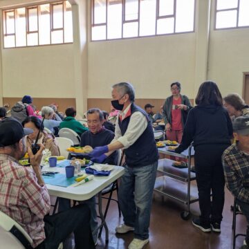 An older man passes out plates of food to older adults seated at a table.