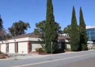 Fenced off one-story commercial building with trees around the lot with downtown Sunnyvale in background