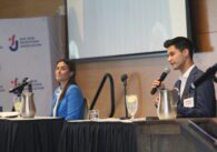 Two people sitting at a dais in front of microphones take turns speaking.