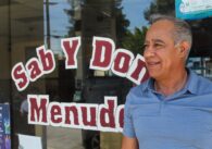 A man standing in front of a store front smiling and looking off camera.