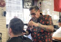Man with slicked back hair and goatee wearing printed shirt combs and trims a customer's hair in a well decorated barbers shop