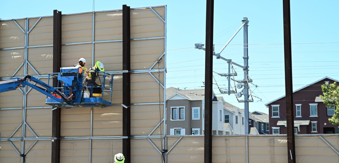 Two workers on a crane are working to install a panelled wall, with apartment buildings visible through a gap in the wall
