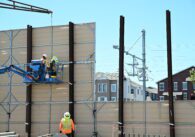 Two workers on a crane are working to install a panelled wall, with apartment buildings visible through a gap in the wall