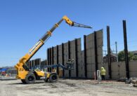 A crane lowering large panels against I-beams in the ground, constructing a wall between an open construction field and a residential neighborhood