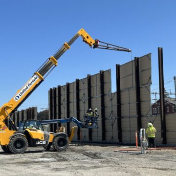 A crane lowering large panels against I-beams in the ground, constructing a wall between an open construction field and a residential neighborhood