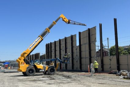A crane lowering large panels against I-beams in the ground, constructing a wall between an open construction field and a residential neighborhood
