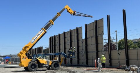 A crane lowering large panels against I-beams in the ground, constructing a wall between an open construction field and a residential neighborhood