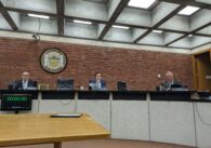 Three men, two older and one young, sit behind a dais in suits