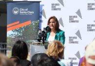 Fair skinned woman with long brunette hair wearing a teal blazer and white shirt speaks at a podium in front of a seated audience, with Bay Area Host Committee and City of Santa Clara banners behind her