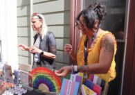 Two women standing behind table at an outdoor venue with open rainbow fan, white and rainbow rubber ducks, multiple resource pamphlets and signs in multiple pride flags