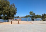 A wooden sign that reads "Almaden Lake Park Terrace Lawn Ampitheater" stands in front of a lake and trees