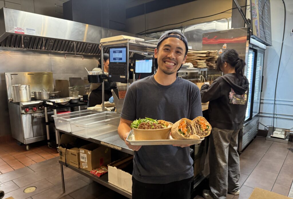 A man in a restaurant kitchen holding a bowl of noodles and a banh mi sandwich