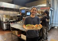 A man in a restaurant kitchen holding a bowl of noodles and a banh mi sandwich