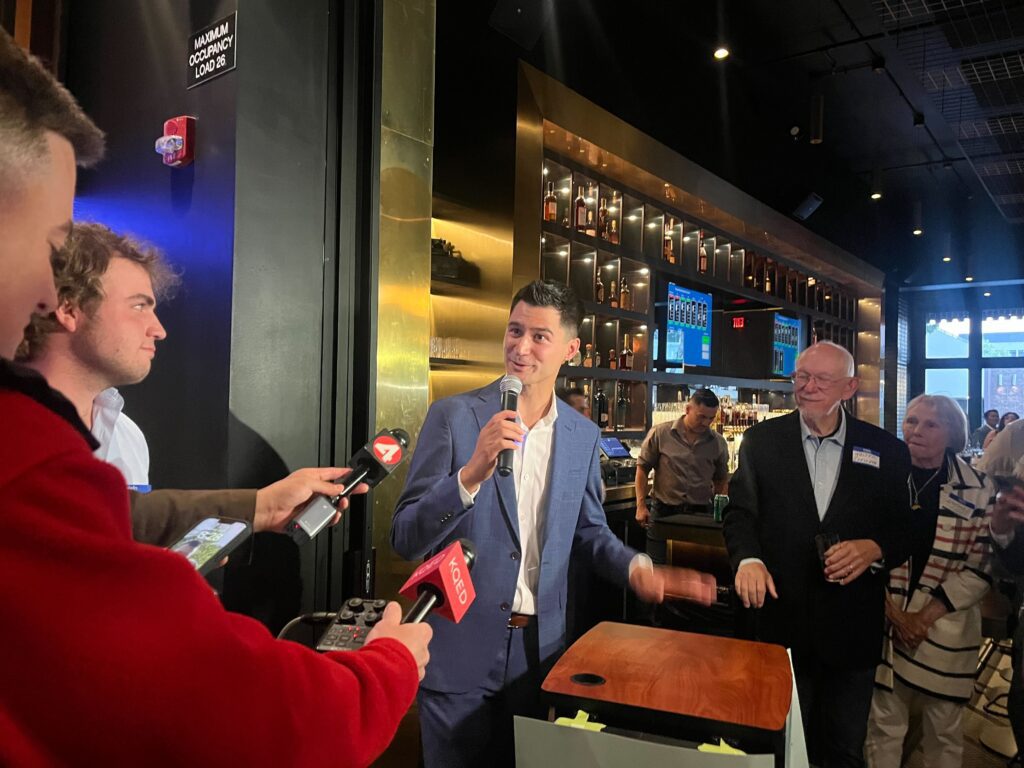 A man in a suit speaking into microphones inside a bar