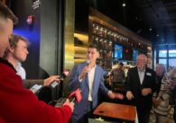 A man in a suit speaking into microphones inside a bar