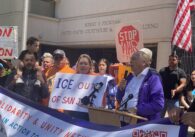 A man speaks into a microphone at a podium with people behind him holding protest signs