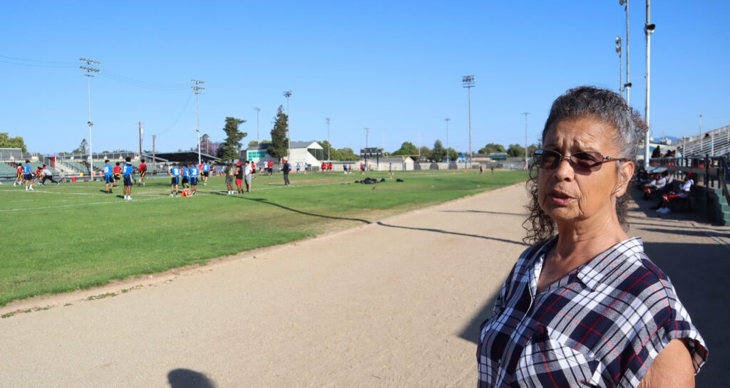 Photo of an older woman with curly white and brown hair wearing a plaid t-shirt and sunglasses stands on a dirt track at an athletic field. Kids are playing flag football on the field in front of her.