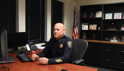 Photo of a bald white man in a police uniform speaking while sitting at a desk, with an American flag and shelves of memorabilia behind him