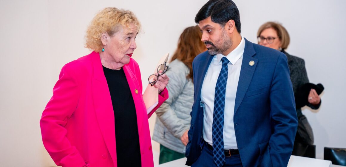 A woman in a pink blazer speaks to a man in a blue suit