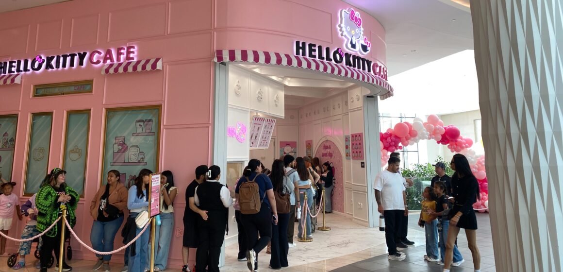 Photo of a bright pink cafe facade with a light up sign of a cute white cat character with a bow, and the words "Hello Kitty Cafe" with a group of people lined up outside