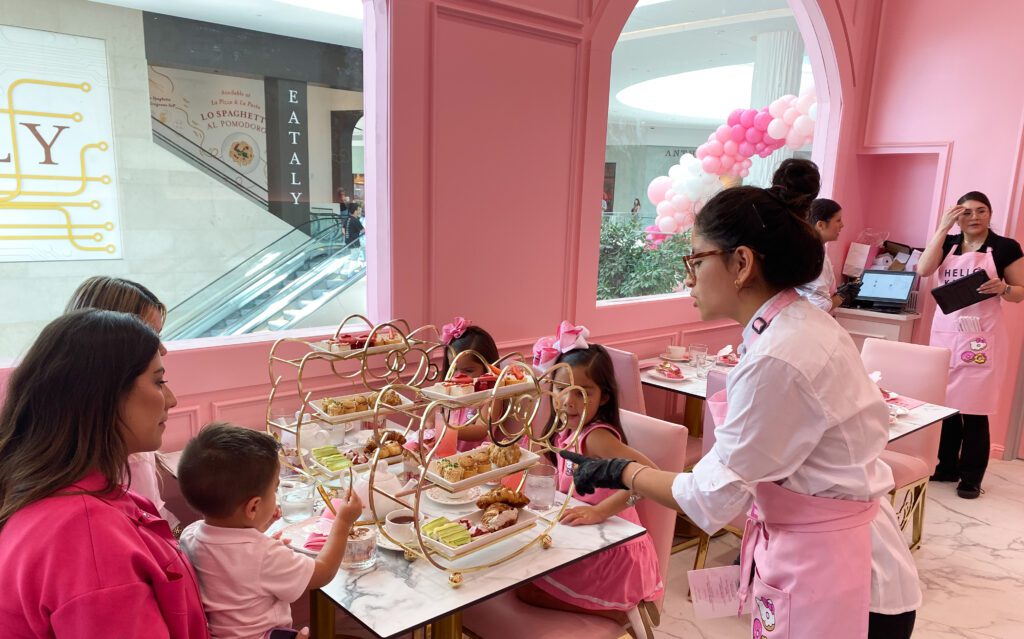 Family of five people — two women, two young girls, and one baby boy — dressed in pink in a pink room with a waitress in a white blouse and pink apron serves out a Hello Kitty shaped pastry holder