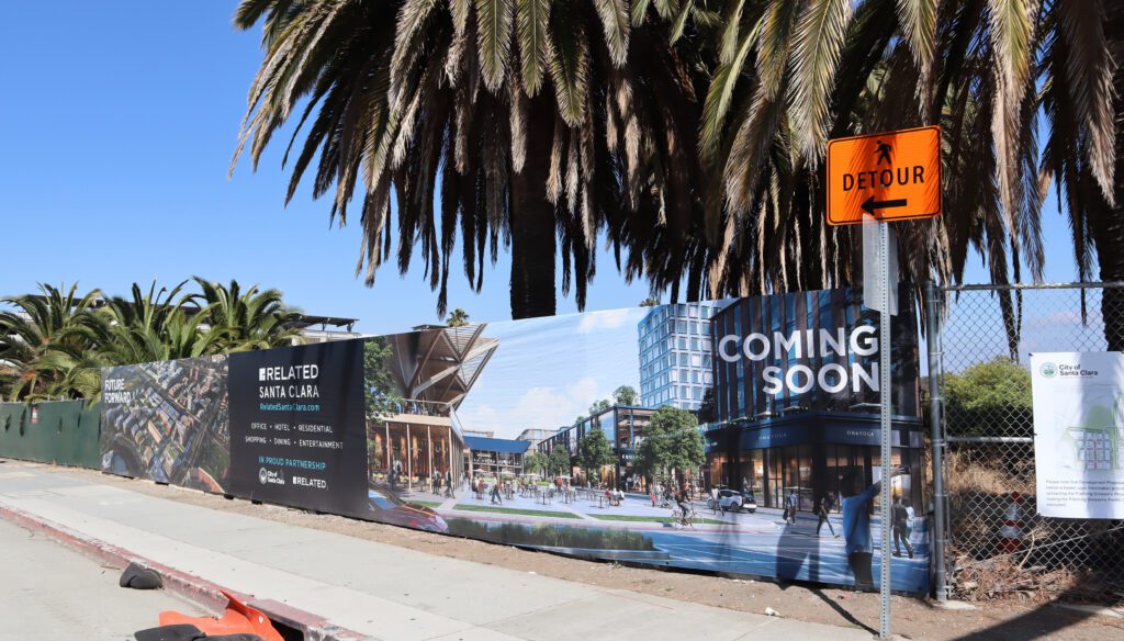 Photo of a fence blocking off a construction area with a pedestrian detour sign, and an advertisement on the fence reading "Coming soon" and "Related Santa Clara: office, hotel, residential, shopping, dining, entertainment." It also says "in proud partnership" with the city of Santa Clara logo.