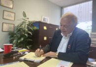 A man in a blazer writes on a pad of paper on a desk in his office