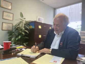 Một người đàn ông mặc áo khoác đang viết trên một tập giấy trên bàn làm việc trong văn phòng của mình