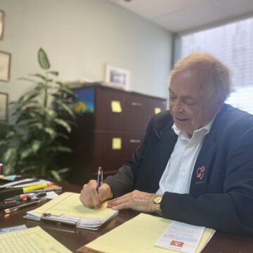 A man in a blazer writes on a pad of paper on a desk in his office