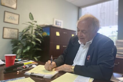 A man in a blazer writes on a pad of paper on a desk in his office