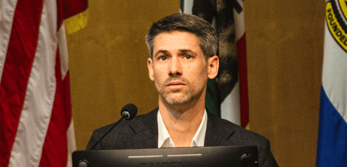 A man in a suit sits behind a computer and microphone at a government meeting
