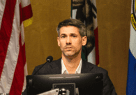 A man in a suit sits behind a computer and microphone at a government meeting