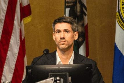 A man in a suit sits behind a computer and microphone at a government meeting