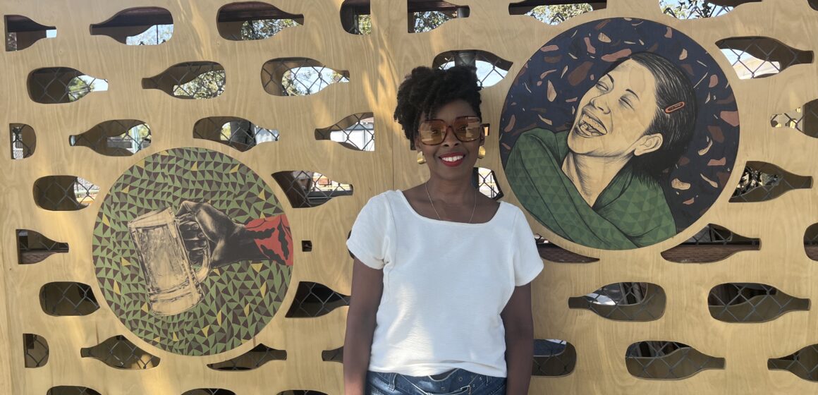 A woman stands in front of art she made displayed on a wall outside
