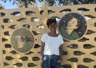 A woman stands in front of art she made displayed on a wall outside