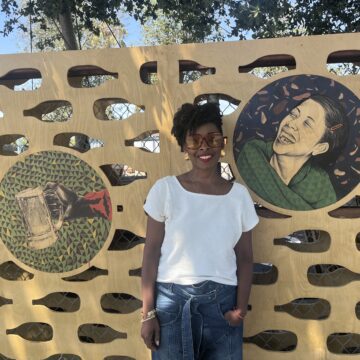 A woman stands in front of art she made displayed on a wall outside