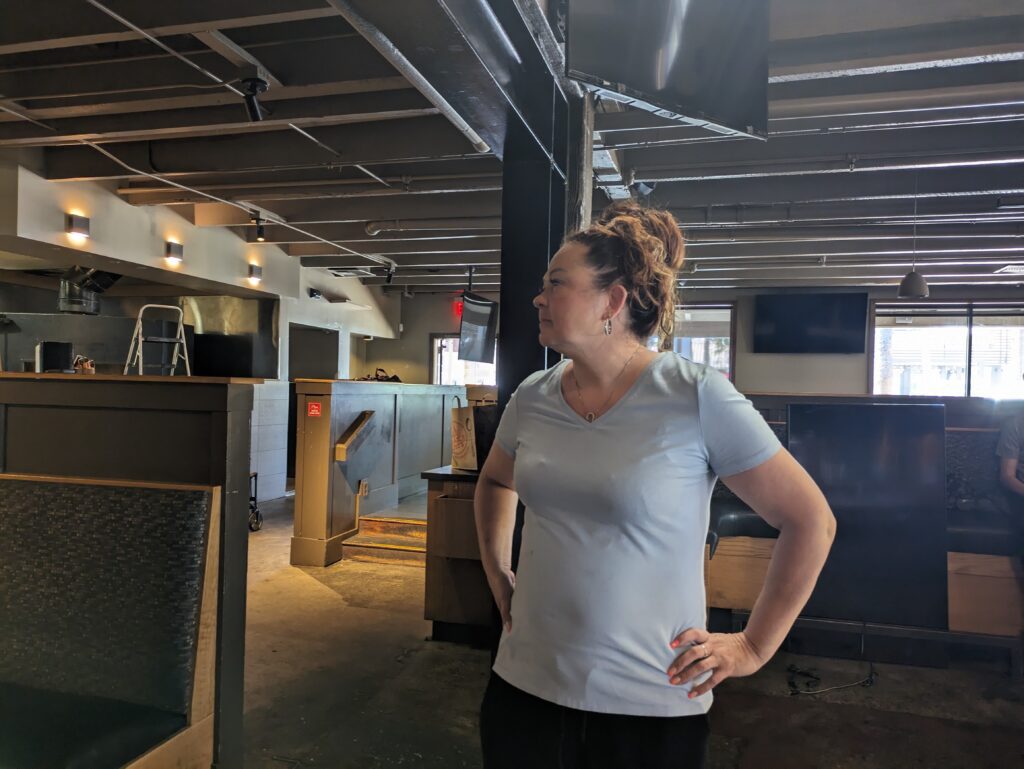 A middle-aged woman with her brown hair in a bun stands in an empty restaurant with her hands on her hips