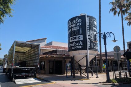 The outside of a restaurant with a moving truck and large water tower with words related to beer on it.