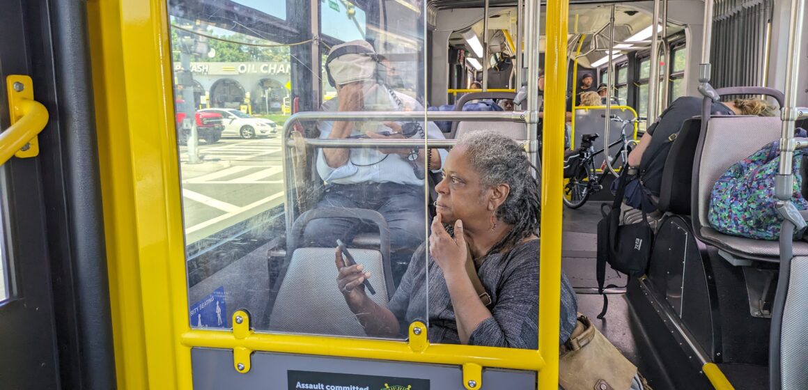 An older woman sits on a public bus with her phone in her hand and bag over her shoulder looking out the window