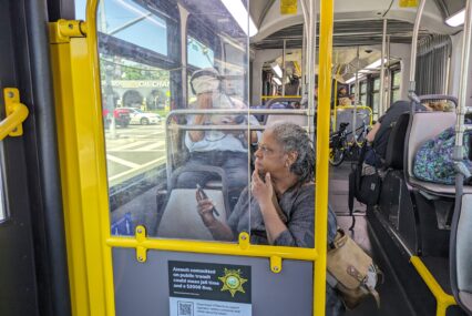 An older woman sits on a public bus with her phone in her hand and bag over her shoulder looking out the window