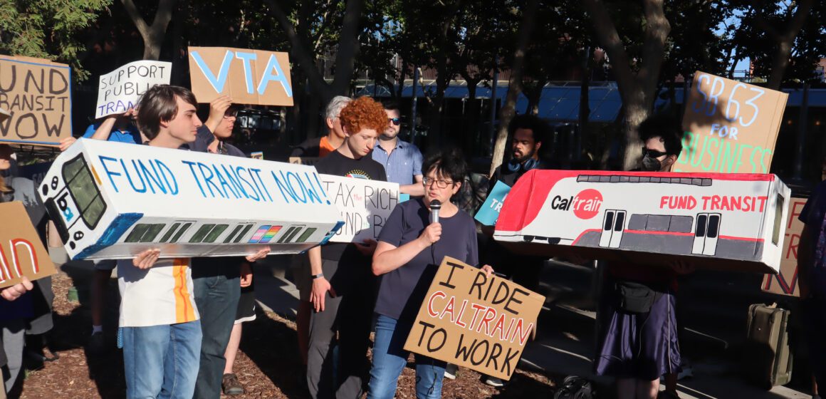 Photo of about 30 people standing outside a building. Nearly everyone is holding signs with slogans such as "Fund transit now" and "Support Public Transit." A shorter woman with short black hair and glasses is holding a sign reading "I ride Caltrain to work" and is speaking into a microphone