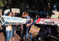 Photo of about 30 people standing outside a building. Nearly everyone is holding signs with slogans such as "Fund transit now" and "Support Public Transit." A shorter woman with short black hair and glasses is holding a sign reading "I ride Caltrain to work" and is speaking into a microphone